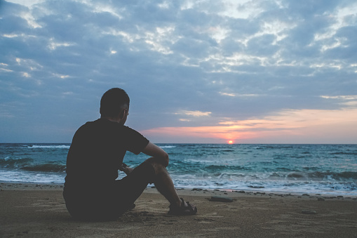 rueful, sorrowful, mournful, sorry, unhappy, wistful alone man looking at the sunset on a beach. concept of sadness loneliness longing and depression. guy at the background of a beautiful sunrise