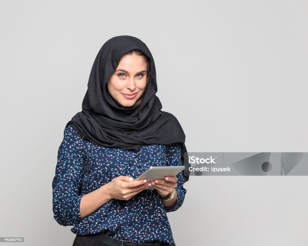 Cheerful young muslim woman holding digital tablet Portrait of cheerful young woman holding digital tablet. Businesswoman in hijab looking at camera against grey background. 20-29 Years Stock Photo
