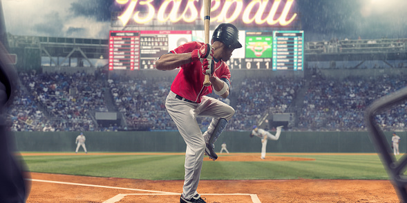 Rear view of a professional baseball batter holding bat upwards with front leg raised, ready to strike the baseball just been thrown by the pitcher. The batter stands in front of a video score board and generic neon sign in a packed stadium, during a night game with light rain