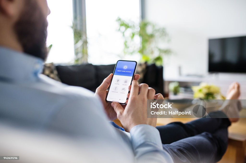 A man holding a smartphone with smart home screen. An unrecognizable man holding a smartphone with smart home control system. Home Automation Stock Photo