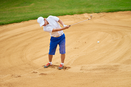 Asian player swinging club in sand trap of a course