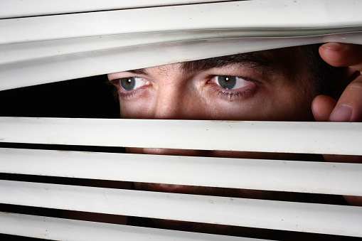 Danger man looking through blinds waiting for someone. Image composition is very dark to represent the dreary atmosphere.