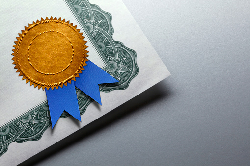 A close up of a gold seal adorned with a blue ribbon is attached to the corner of a certificate of achievement which rests on top of a gray background which provides ample room for copy or text.