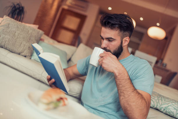 istock Young man is sitting on a sofa and reading a book 942722346