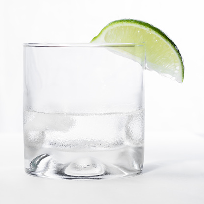 close up of a short glass with a clear liquid and a green lime on the rim isolated on a white background
