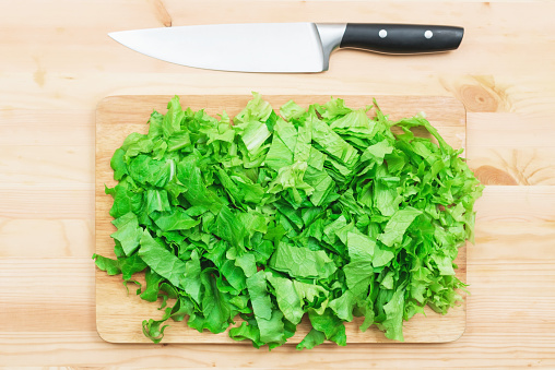Close-up on a wooden table is a wooden cutting board on which lies chopped lettuce leaves and next to a large cutting knife. The concept of self-cooking vegetarian food at home. Healthy lifestyle.