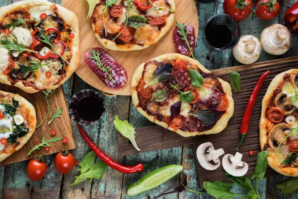 Flatlay of pizza party. Pizzas in abundance with raw vegetables, salami, mushrooms and red wine on dark oak chopping boards over blue rustic background overhead view
