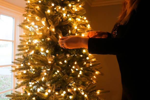 istock Blond woman dressed in black decorating christmas tree with lights by bay window 942777494