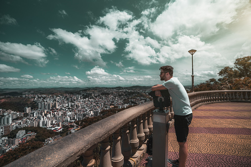 Thoughtful bearded hipster guy in a white t-shirt, black shorts, and sunglasses is leaning on the monocular while standing in the observation area on the top of the city and looking at urban landscape