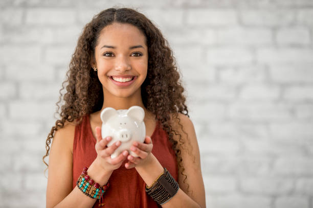 Saving Up Money A teenage girl with long curly hair is indoors. She is smiling at the camera while holding a piggy bank. She is saving money for a big purchase. financial wellbeing stock pictures, royalty-free photos & images