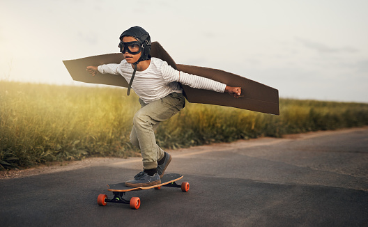Shot of a young boy pretending to fly with a pair of cardboard wings while riding a skateboard outside