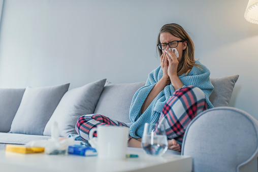 Sick woman with a headache sitting on a sofa at home wrapped in blanket during the day. Sickness, seasonal virus problem concept. Woman being sick having flu lying on sofa, Blowing or sneezing nose