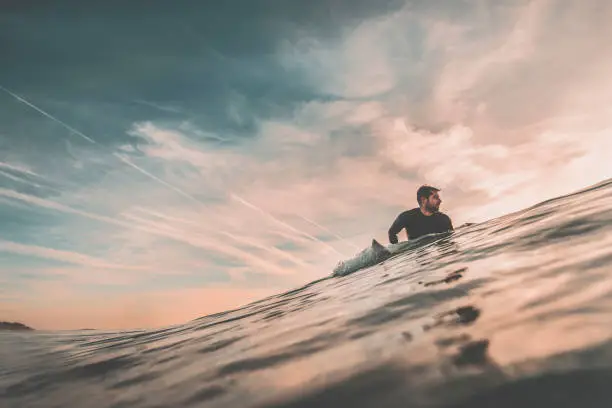 Young man waits in the sea, near the coast, on his surfboard, a big wave at sunset. Extreme water sports and outdoor active lifestyle. Vintage filter with soft style