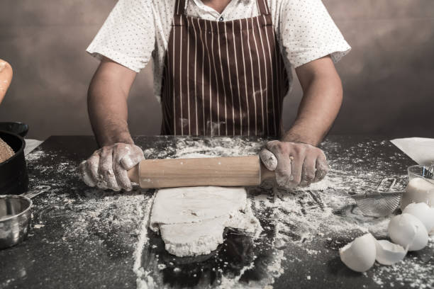 istock Man preparing buns at table in bakery, Man sprinkling flour over fresh dough on kitchen table 942942288