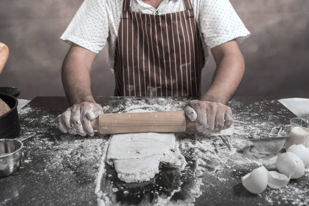 istock Man preparing buns at table in bakery, Man sprinkling flour over fresh dough on kitchen table 942946548