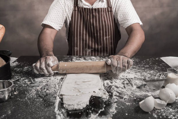 istock Man preparing buns at table in bakery, Man sprinkling flour over fresh dough on kitchen table 942949166