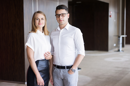 Portrait of confident business colleagues posing in office building. Young Caucasian businesswoman and businessman wearing glasses standing together and smiling. Team and cooperation concept
