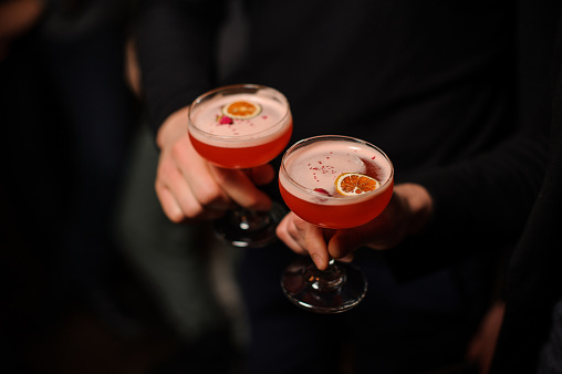 Two people dressed in black holding cocktail glasses filled with sweet alcoholic drink with orange