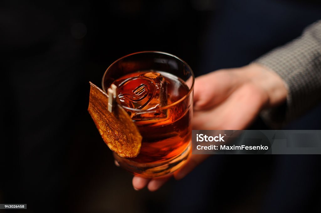 Male hand holding a glass of whiskey with ice Male hand holding a glass of whiskey with ice cube on the dark blurred background Alcohol - Drink Stock Photo