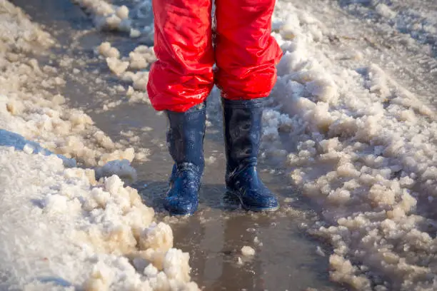 Kid legs in blue rainboots standing in the ice puddle with melting snow at sunny spring day, outdoors