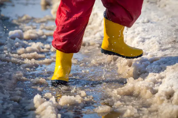 Kid legs in yellow rainboots running in the ice puddle with melting snow at sunny spring day, outdoors