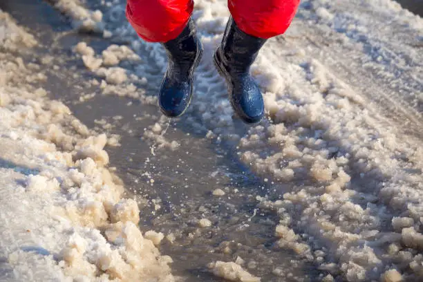 Kid in blue rainboots jumping in the ice puddle with melting snow at sunny spring day, outdoors