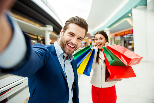 It's shopping and fun time. Cute selfie portrait of cheerful  successful happy young lovely couple holding  colored shopping bags and laughing in mall at holiday