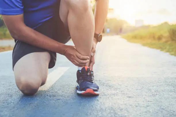 Photo of Injury from workout concept : Asian man use hands hold on his ankle while running on road in the park. Focus on ankle.