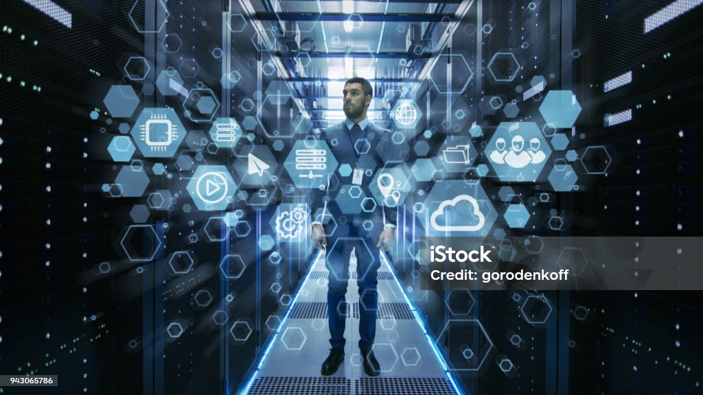 Curios IT Engineer Standing in the Middle of a Working Data Center Server Room. Cloud and Internet Icon Visualization in the Foreground. Cloud Computing Stock Photo
