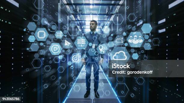 Curios It Engineer Standing In The Middle Of A Working Data Center Server Room Cloud And Internet Icon Visualization In The Foreground Stock Photo - Download Image Now