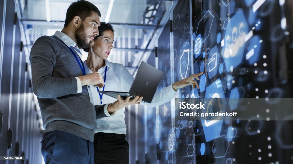 Male IT Specialist Holds Laptop and Discusses Work with Female Server Technician. They're Standing in Data Center, Rack Server Cabinet with Cloud Server Icon and Visualization. Technology Stock Photo
