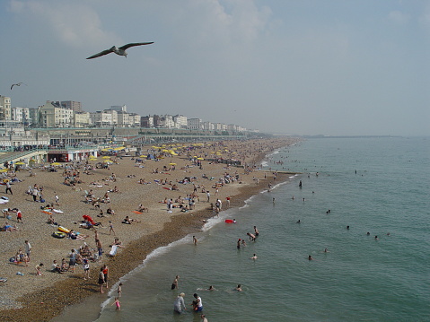 View from pier of the Brighton (UK) beach on a hot sunny day: people of different ages and cultures enjoying same thing - sunshine and the sea. Brighton seafront, blue sky, seagulls fly.