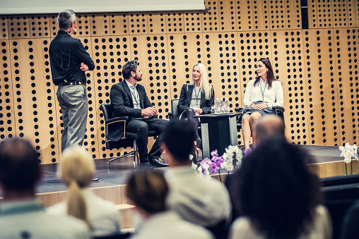 Four people on stage participating in a round table, one is asking questions while others are answering. Rear view of visible heads of audience members.