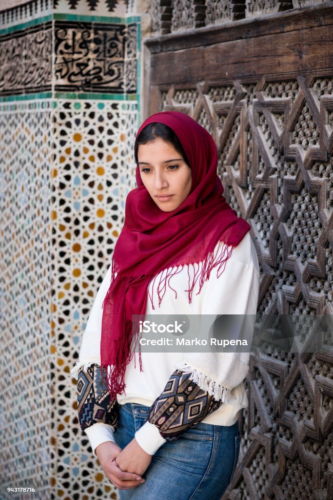 Nostalgic muslim woman in traditional clothing with headscarf and jeans Nostalgic muslim woman in traditional clothing with red hijab and jeans in front of traditional arabesque decorated wall Adult Stock Photo