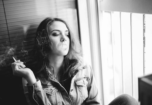 Pensive young girl sitting next to the open door and smoking a cigarette. Black and white.