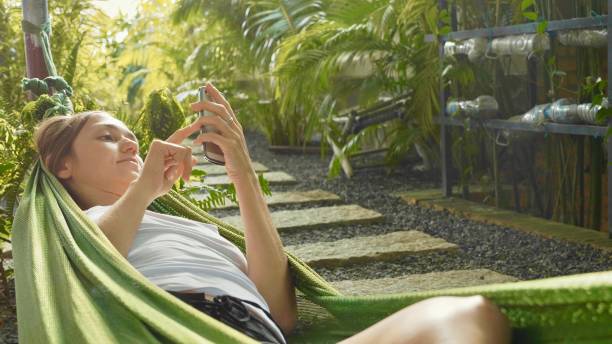 istock Young woman uses a smartphone while lying in a hammock at green garden 943351812