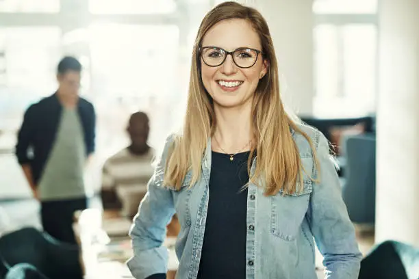 Young designer wearing glasses and smiling confidently while standing in an office with colleagues working behind her