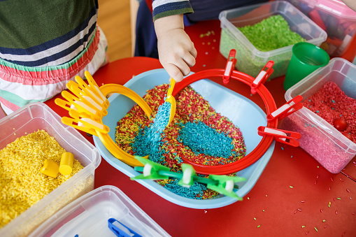 Toddlers playing with sensory bin with colourful rice on red table.
