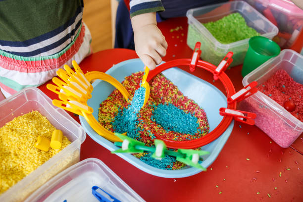 Toddlers playing with sensory bin with colourful rice on red table stock photo