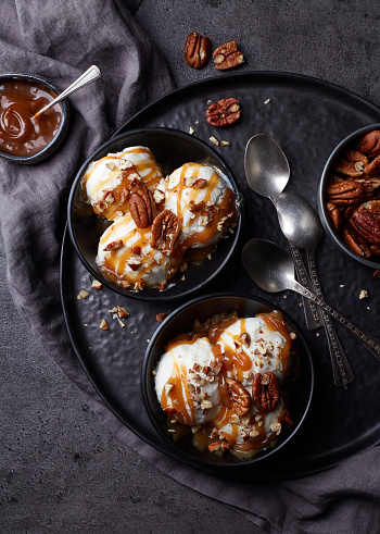 Two black bowls of vanilla ice cream balls with caramel sauce and pecan nuts on dark marble background. Top view