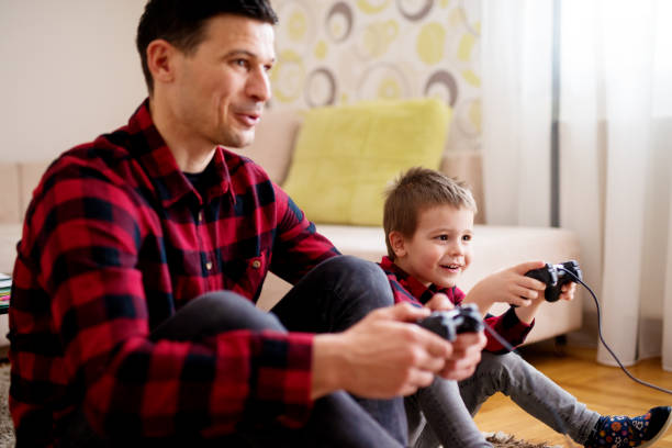 istock Young cheerful excited father and son in the same red shirt playing console games with gamepads in a bright living room. 943513454