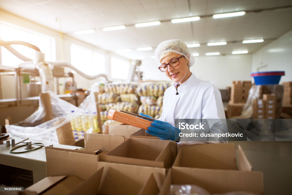 Close up view of a young female pleased worker in sterile cloths packing finished food products in a boxes in food factory storage. Healthcare And Medicine Stock Photo
