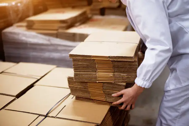 Photo of Close up of young female worker picking up stacks of folded cardboard boxes from a bigger stack in factory storage room.