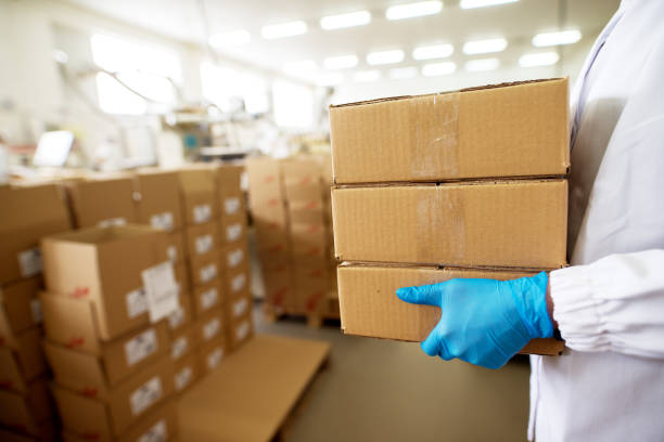 istock Close up view of a worker carrying a stack of packages in brown boxes within production industry storage room while wearing sterile cloths and rubber gloves. 943521836