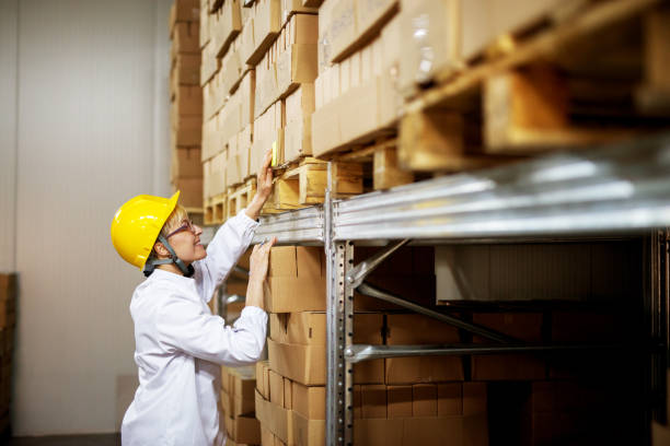 istock Young joyful female worker reading sticker note on a stack of brown boxes on pallets in a factory storage area. 943525152