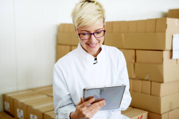 istock Young joyful female technician is smiling while using a tablet and leaning against the big stack of cardboard boxes. 943528524
