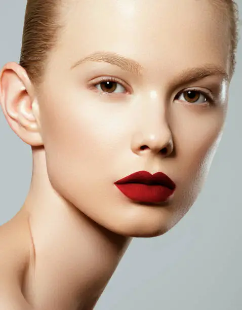 Beauty woman Face. Portrait of beautiful sexy young female with perfect matte facial makeup. Soft healthy skin. Glamorous girl on gray background. Red lips