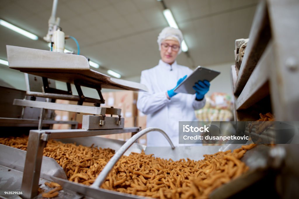 Close up view of a salt stick snacks being processed through a production line in food factory while being inspected by a female young worker with a tablet in a sterile cloth. Food Stock Photo