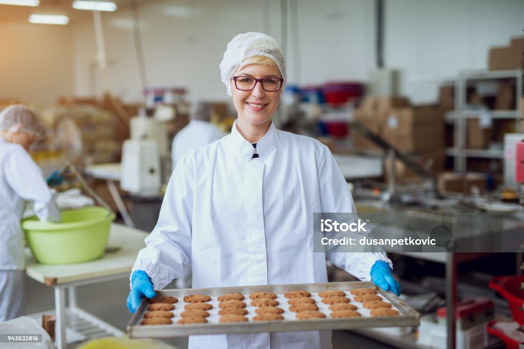 Jeune travailleur féminin heureux en chiffons stériles tenant fraîchement biscuits sur fer-blanc à l’intérieur de l’usine de production d’aliments cuits au four. - Photo de Usine agro-alimentaire libre de droits