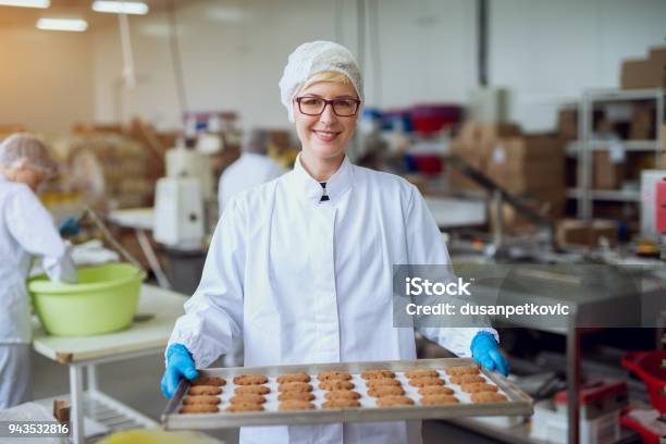 Photo libre de droit de Jeune Travailleur Féminin Heureux En Chiffons Stériles Tenant Fraîchement Biscuits Sur Ferblanc À Lintérieur De Lusine De Production Daliments Cuits Au Four banque d'images et plus d'images libres de droit de Usine agro-alimentaire